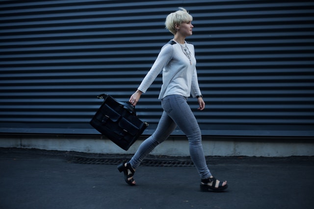 Žena s blond vlasmi, v bielom tričku a sivých nohaviciach s čiernou kabelkou kráča po ulici